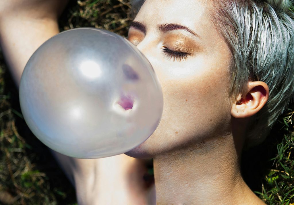 A close-up of a woman blowing a bubble with bubblegum while lying outdoors.