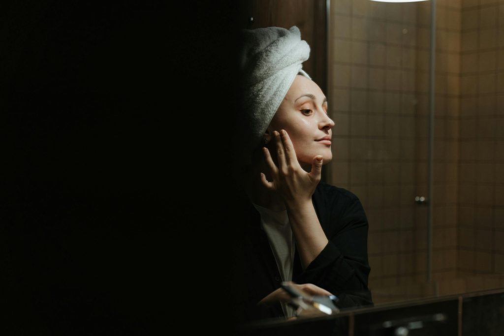 A woman enjoys her skincare routine in a dimly lit bathroom mirror reflection.