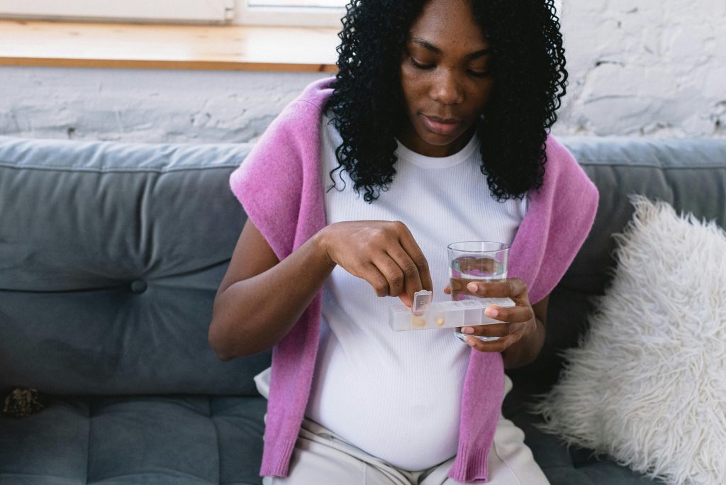 Pregnant woman sitting on a couch taking prenatal vitamins with a glass of water, symbolizing self-care.