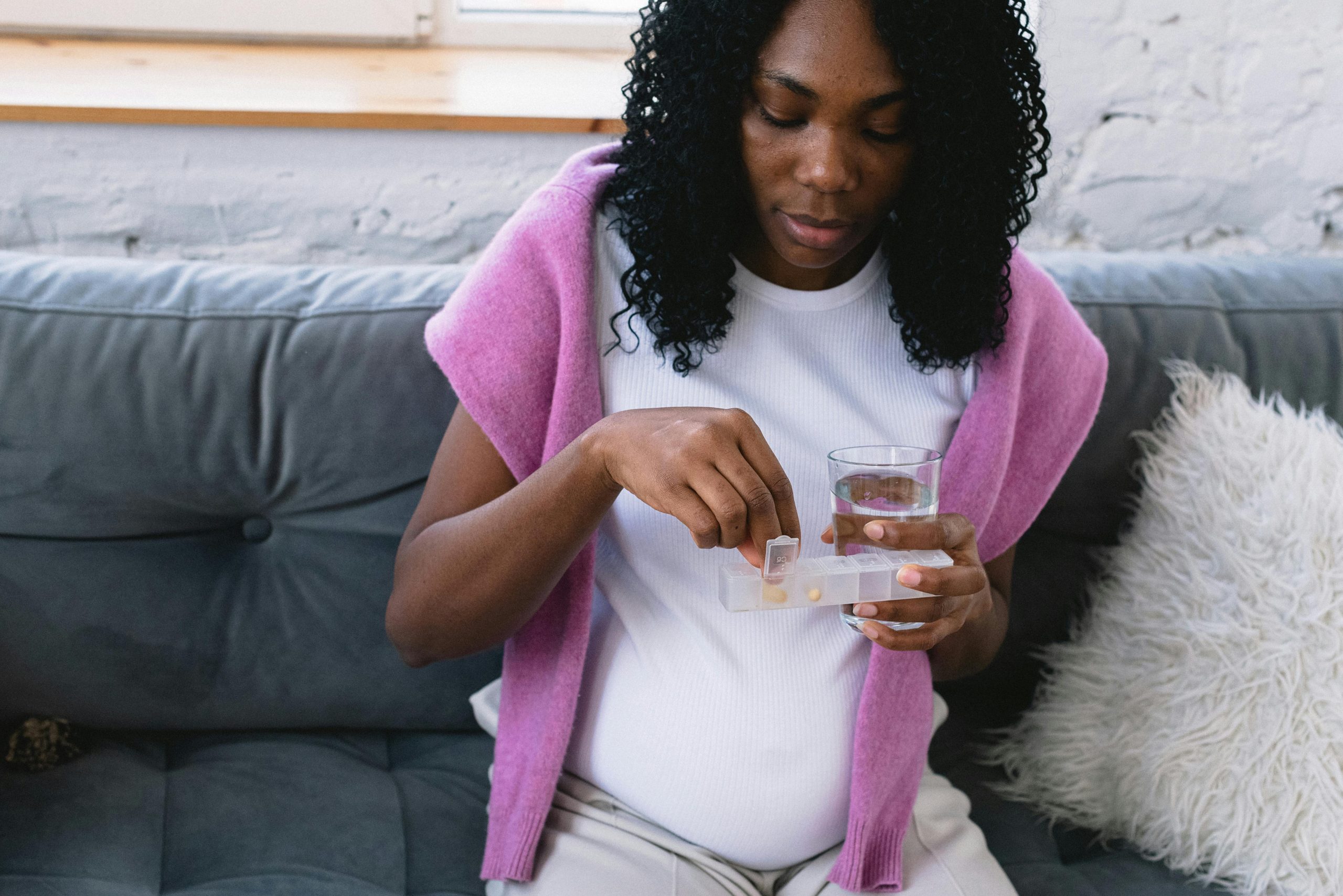 Pregnant woman sitting on a couch taking prenatal vitamins with a glass of water, symbolizing self-care.