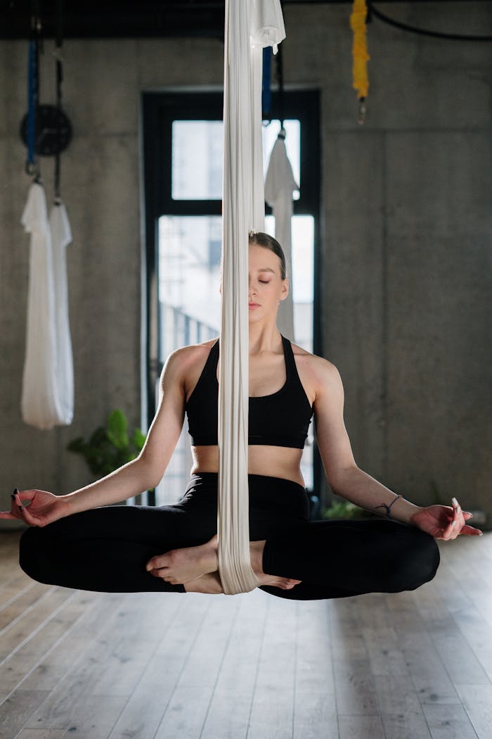 A woman in a yoga studio performing aerial yoga in a suspended lotus pose, focusing on relaxation.