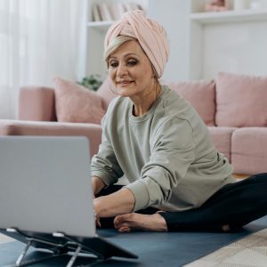 Senior woman using a laptop for an online yoga class, sitting indoors on a yoga mat with a relaxed expression.