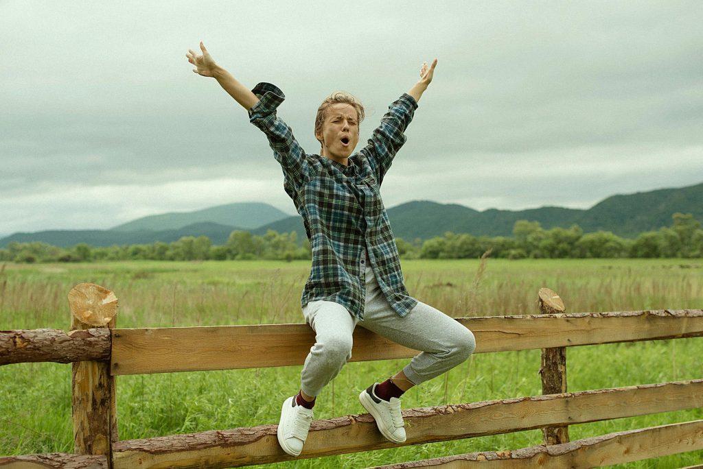 Woman celebrating with arms raised, sitting on a wooden fence in rural landscape.